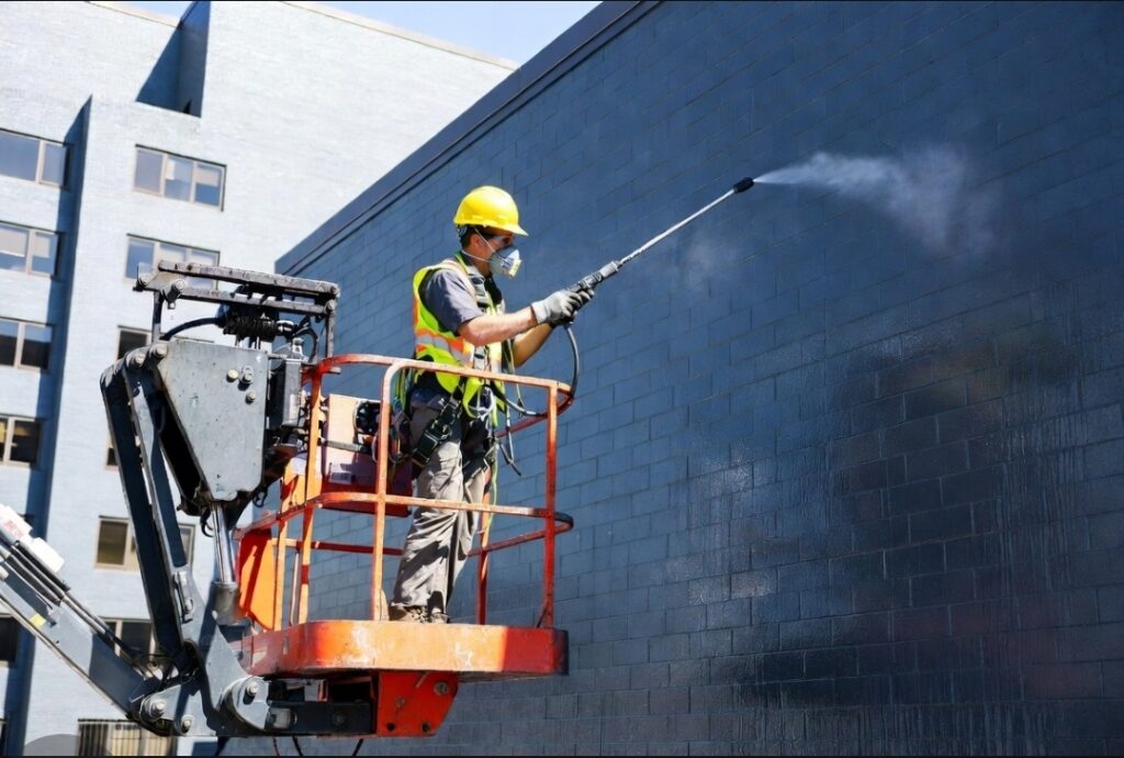 A certified masonry technician from Emerson Masonry Inc in a boom lift applies penetrating sealant for comprehensive masonry waterproofing Indianapolis on a commercial brick building for Emerson Masonry Inc.